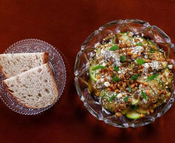 Tigela de vidro com salada contendo fatias de pepino, queijo branco, nozes picadas e ervas verdes, acompanhada por prato com duas fatias de pão integral sobre superfície de madeira.