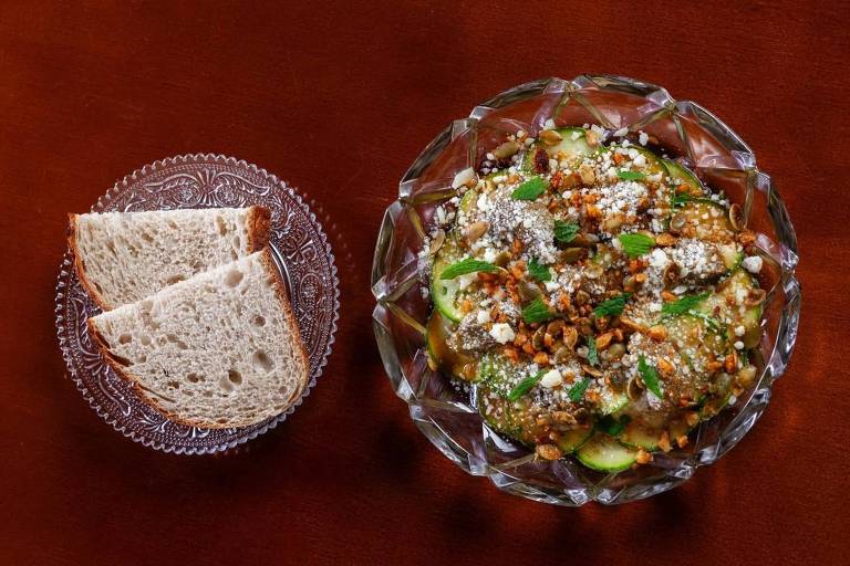 Tigela de vidro com salada contendo fatias de pepino, queijo branco, nozes picadas e ervas verdes, acompanhada por prato com duas fatias de pão integral sobre superfície de madeira.